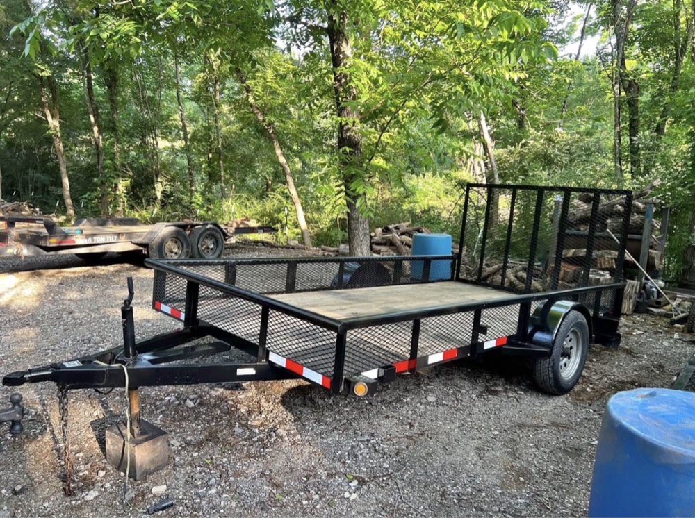 13ft utility trailer, empty, parked in a wooded gravel lot